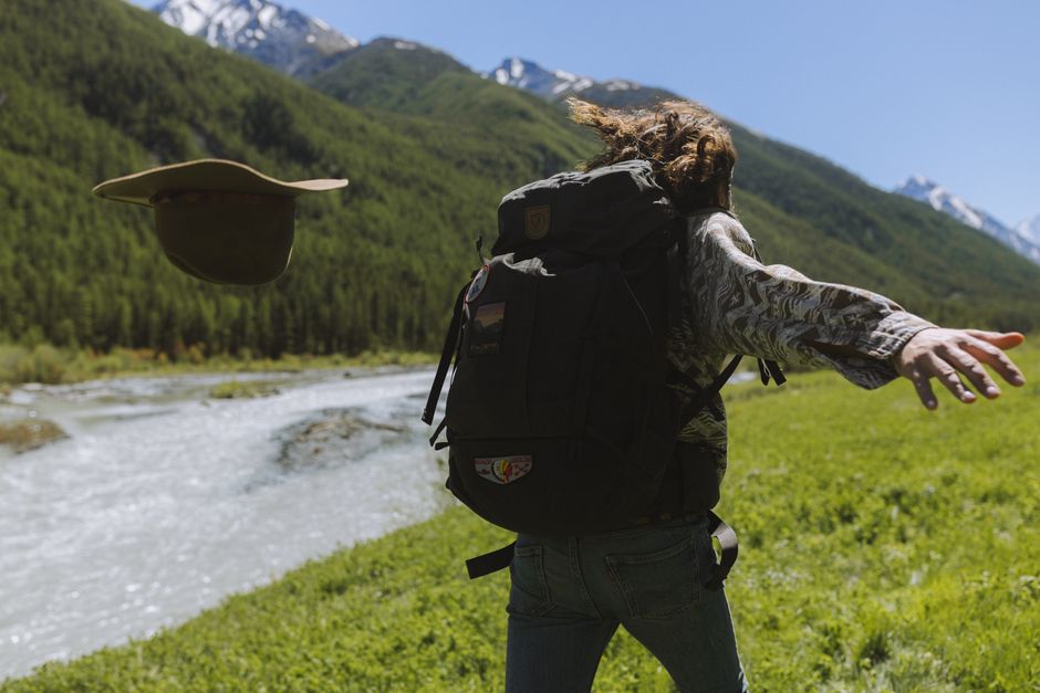 A person with a backpack enjoys the outdoors, tossing a hat near a scenic mountain stream.