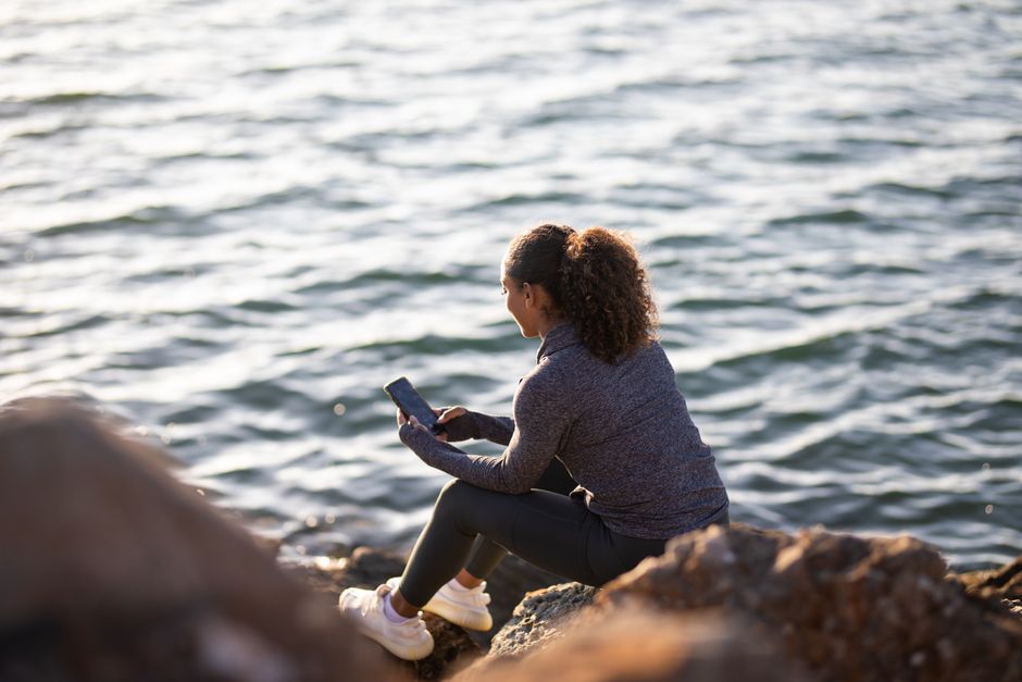 Woman in a sweater sitting on a rocky shore, engaging with smartphone. Serene seaside scene.