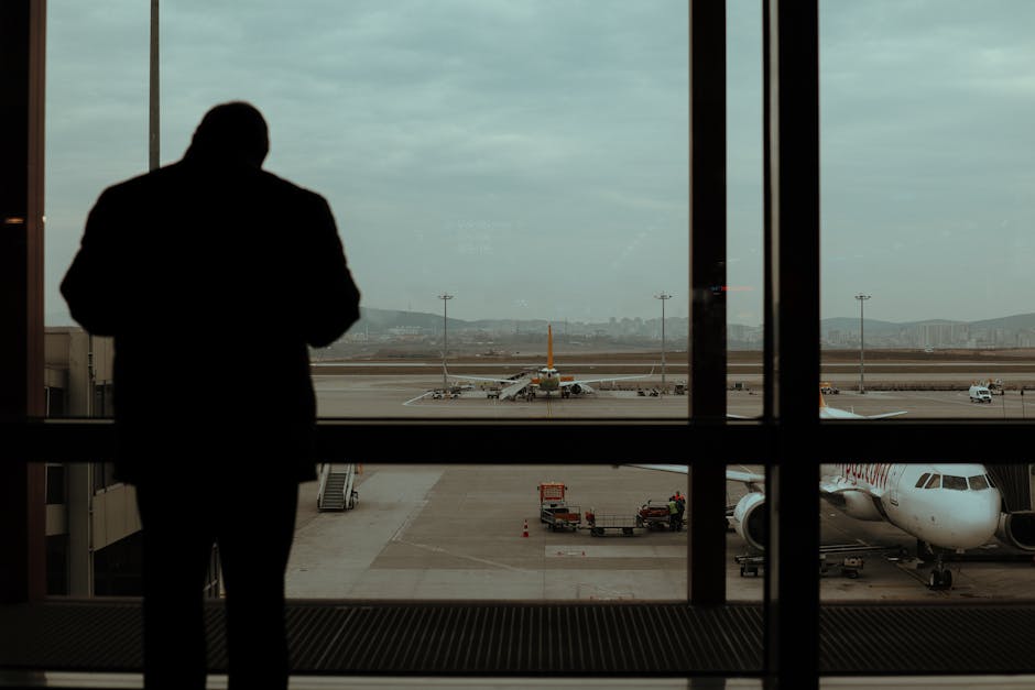 A silhouetted man observes planes and runway through large glass windows at an airport.
