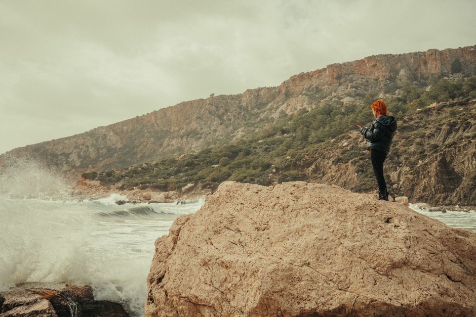 A person stands on a rock by the ocean, surrounded by waves and cliffs.