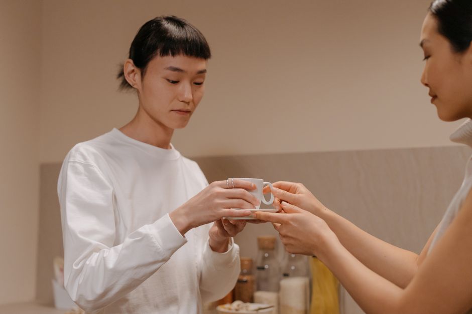 Side view of crop adult Asian woman giving cup of freshly brewed coffee to teenage daughter while standing together in kitchen during breakfast