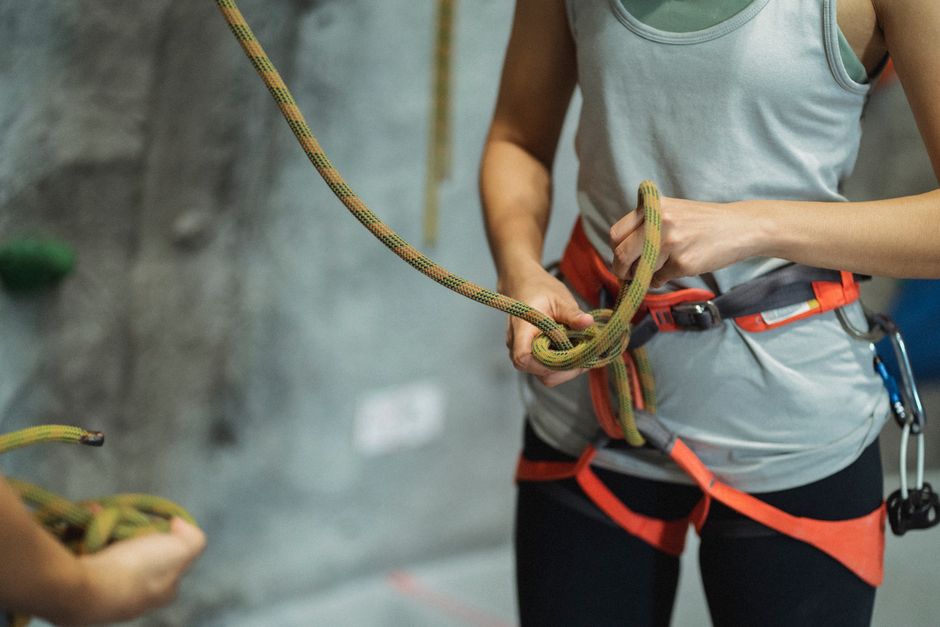 Crop anonymous fit female alpinist in sportswear tying safety rope near crop trainer during practice