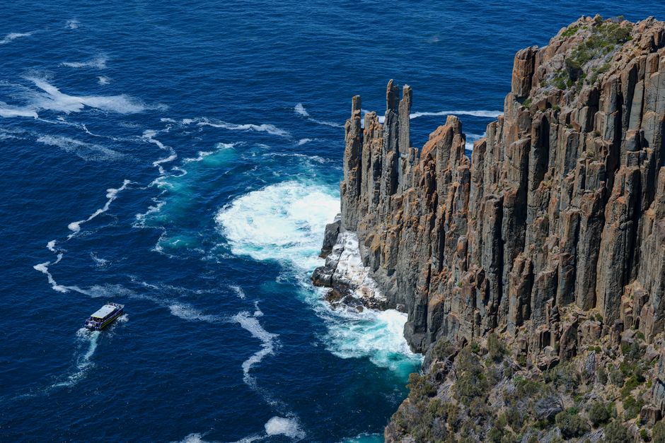 Aerial view of rugged sea cliffs and a boat navigating the blue waters off Tasmania's coast.