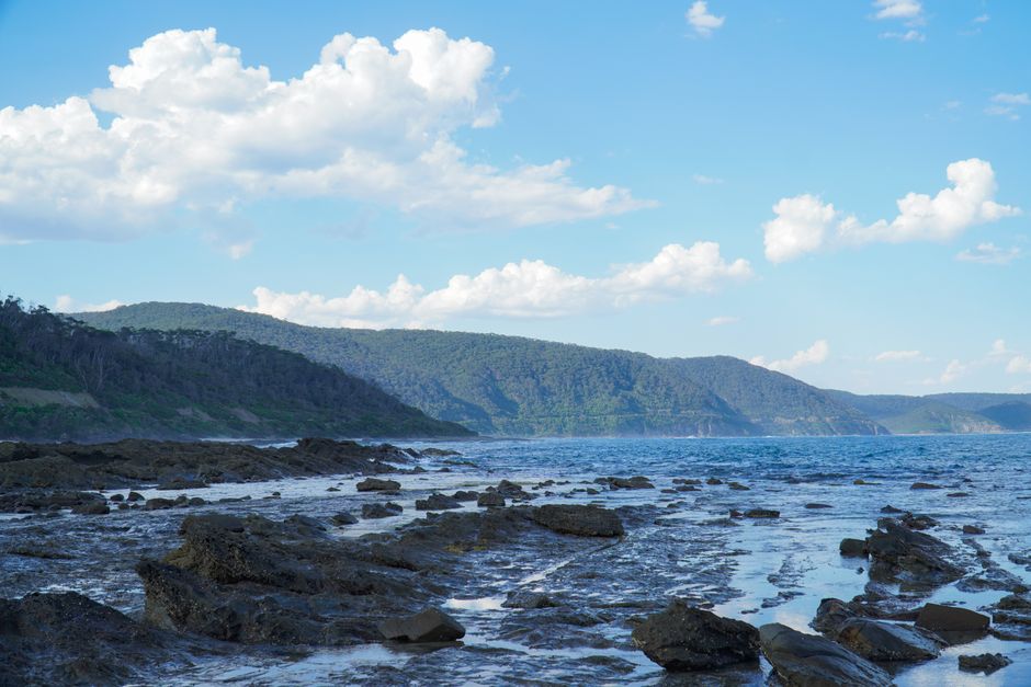 Peaceful coastal scene with rocky shore, ocean, and distant hills under a cloudy sky.