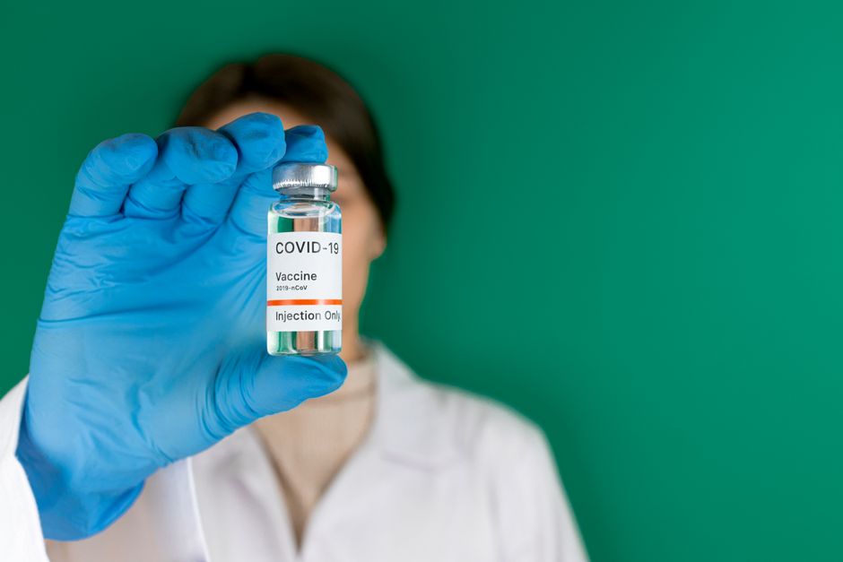 Close-up of a COVID-19 vaccine vial held by a person wearing medical gloves with green background.