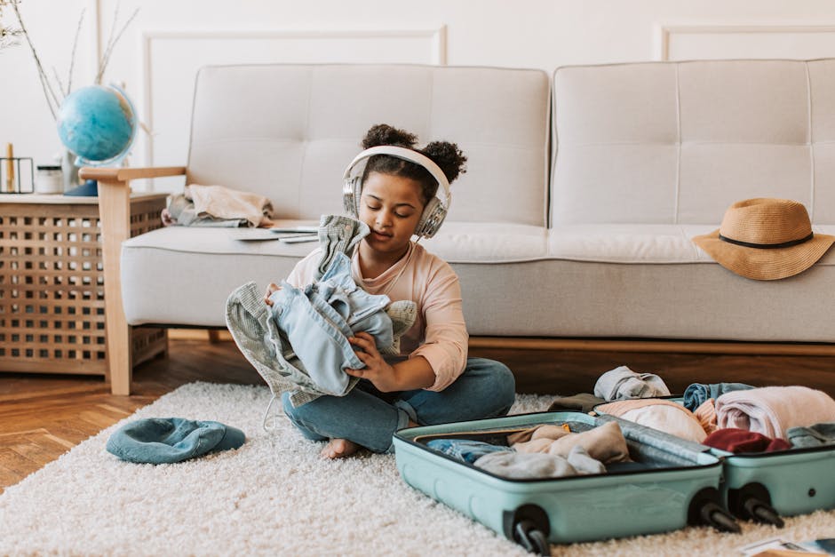 Girl sitting indoors wearing headphones, folding clothes into a suitcase for a trip.